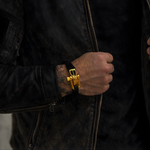 Masculine man wearing a luxury black paracord bracelet with gold shackle with a black leather jacket on a dark background