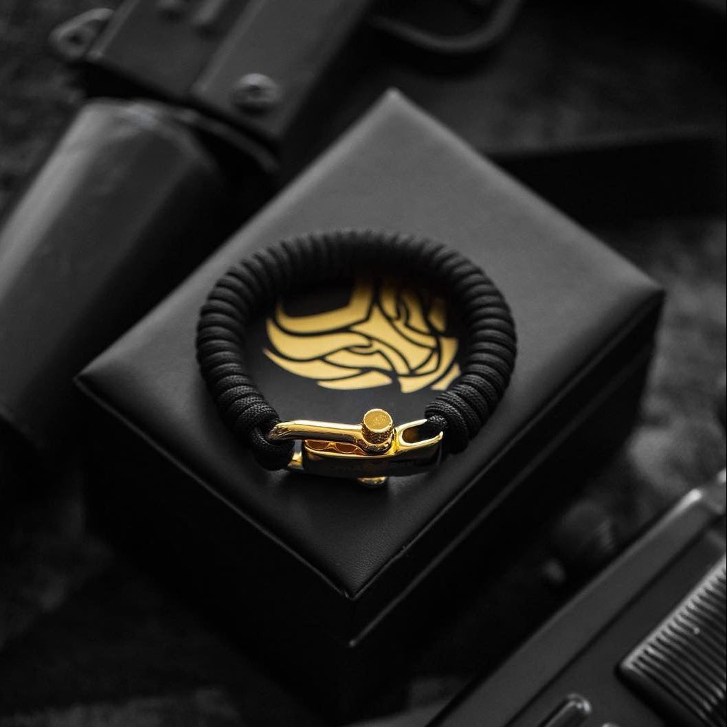 Luxury black paracord bracelet with gold shackle on a black leather case with Alpha Rebel brand logo.